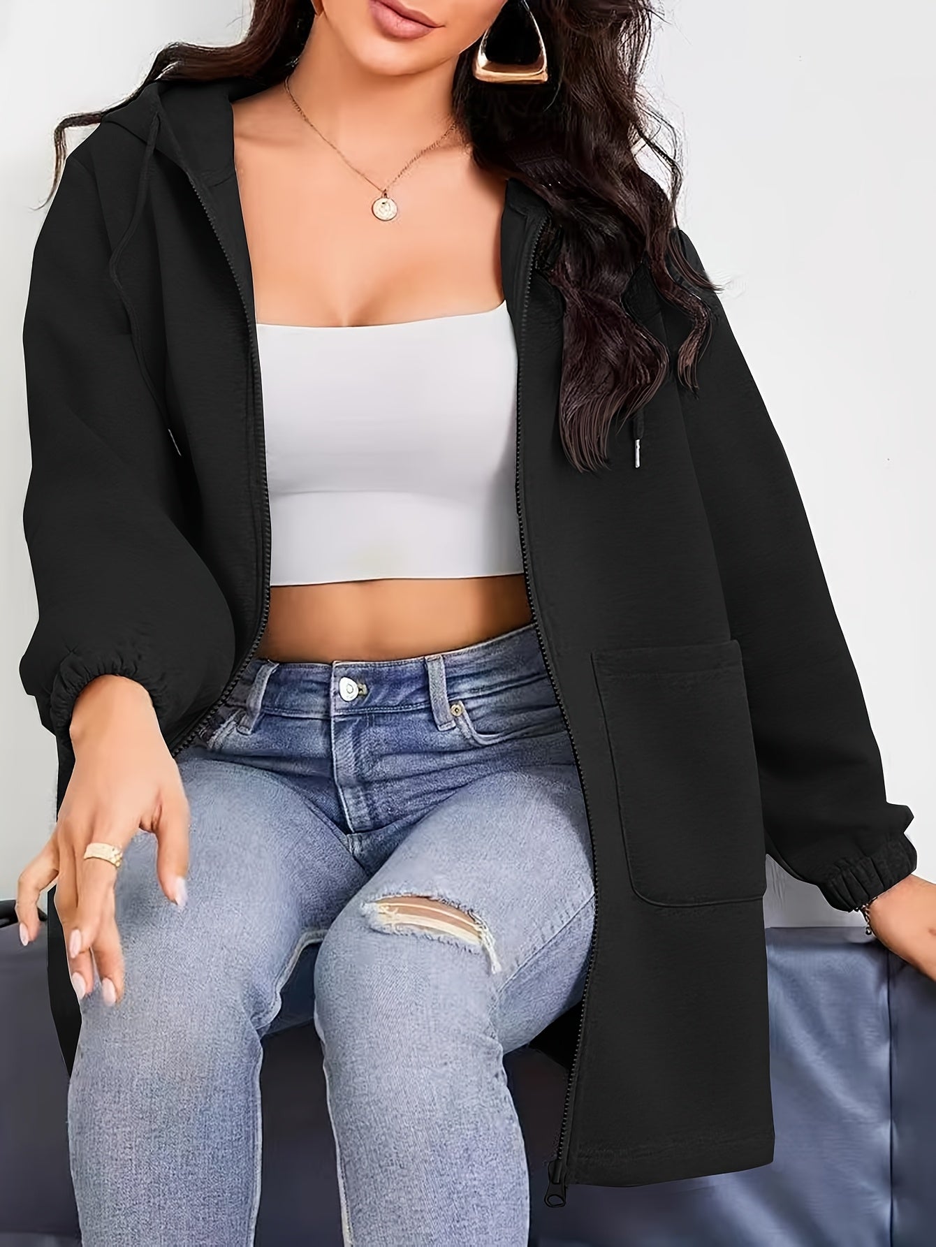 Woman wearing a black women's oversized zip up hoodies, white crop top, and blue jeans on a plain background