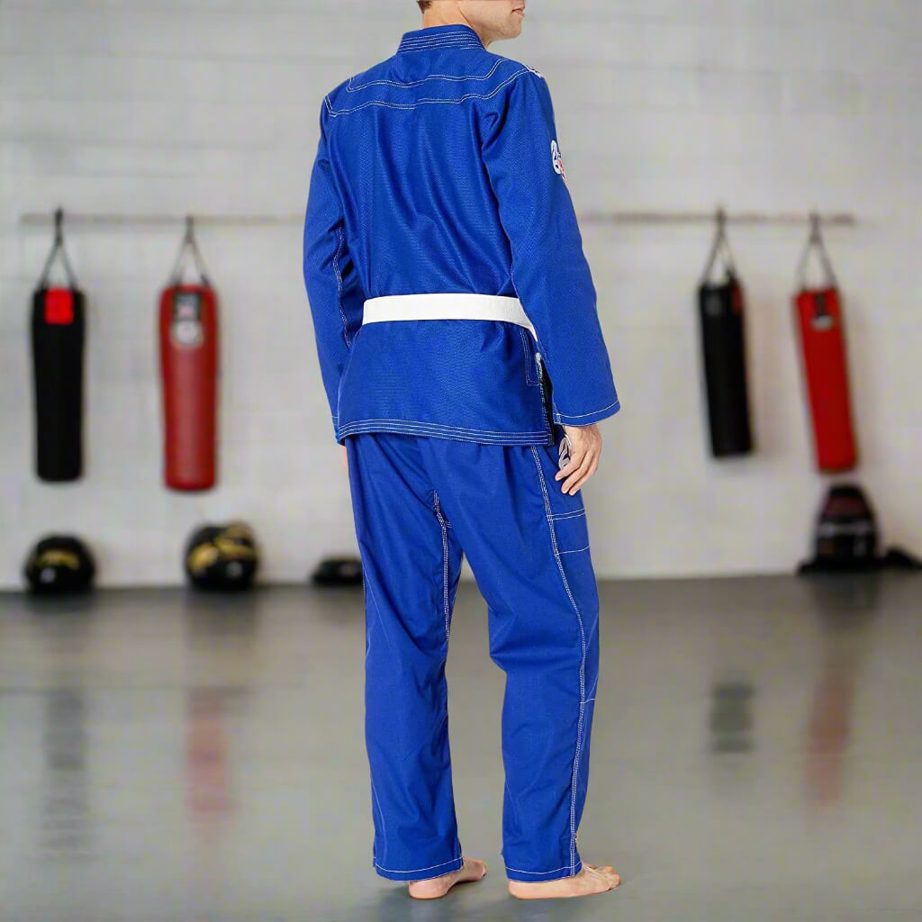 Person wearing a blue martial arts uniform in a gym setting with punching bags in the background.