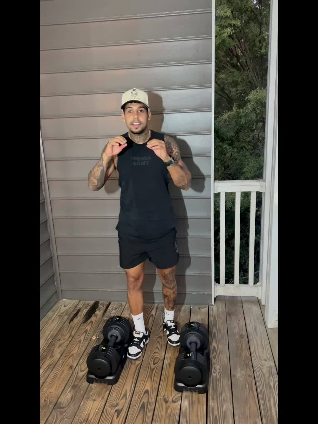 Man standing on a wooden deck with heavy adjustable dumbbell set, wearing a black tank top and shorts.