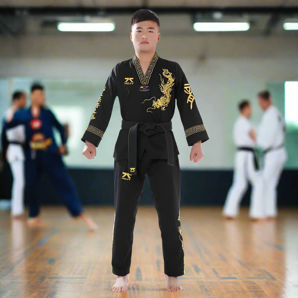 Person wearing a black taekwondo uniform with gold designs in a gym setting.