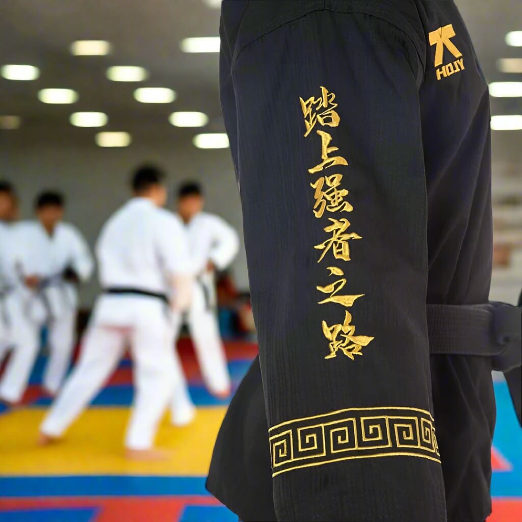 Black martial arts uniform with gold text and patterns on a blurred martial arts training background