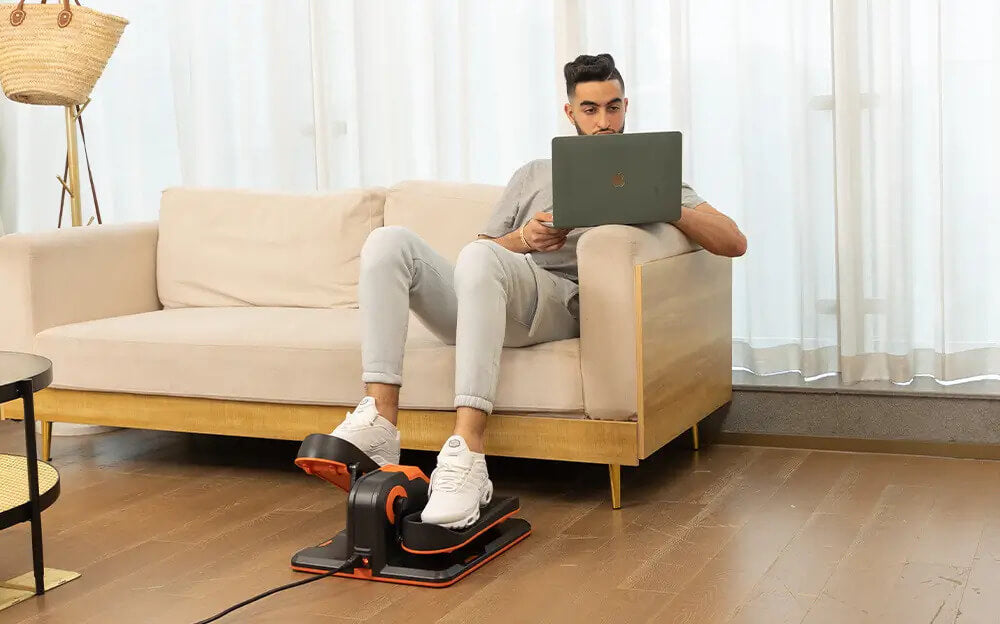 A man working on a laptop while sitting on a sofa in a living room while exercising her legs on an under desk electric elliptical machine sold by 100 Spartans