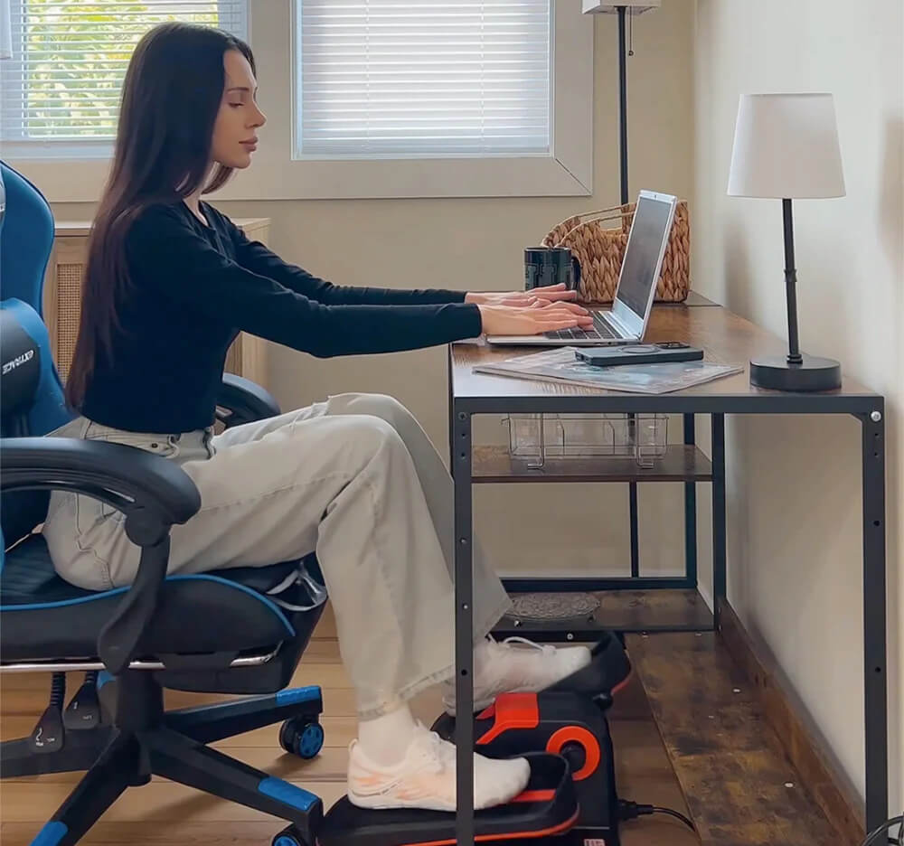 A lady working on a laptop on their work station while exercising her legs on an under desk electric elliptical machine sold by 100 Spartans