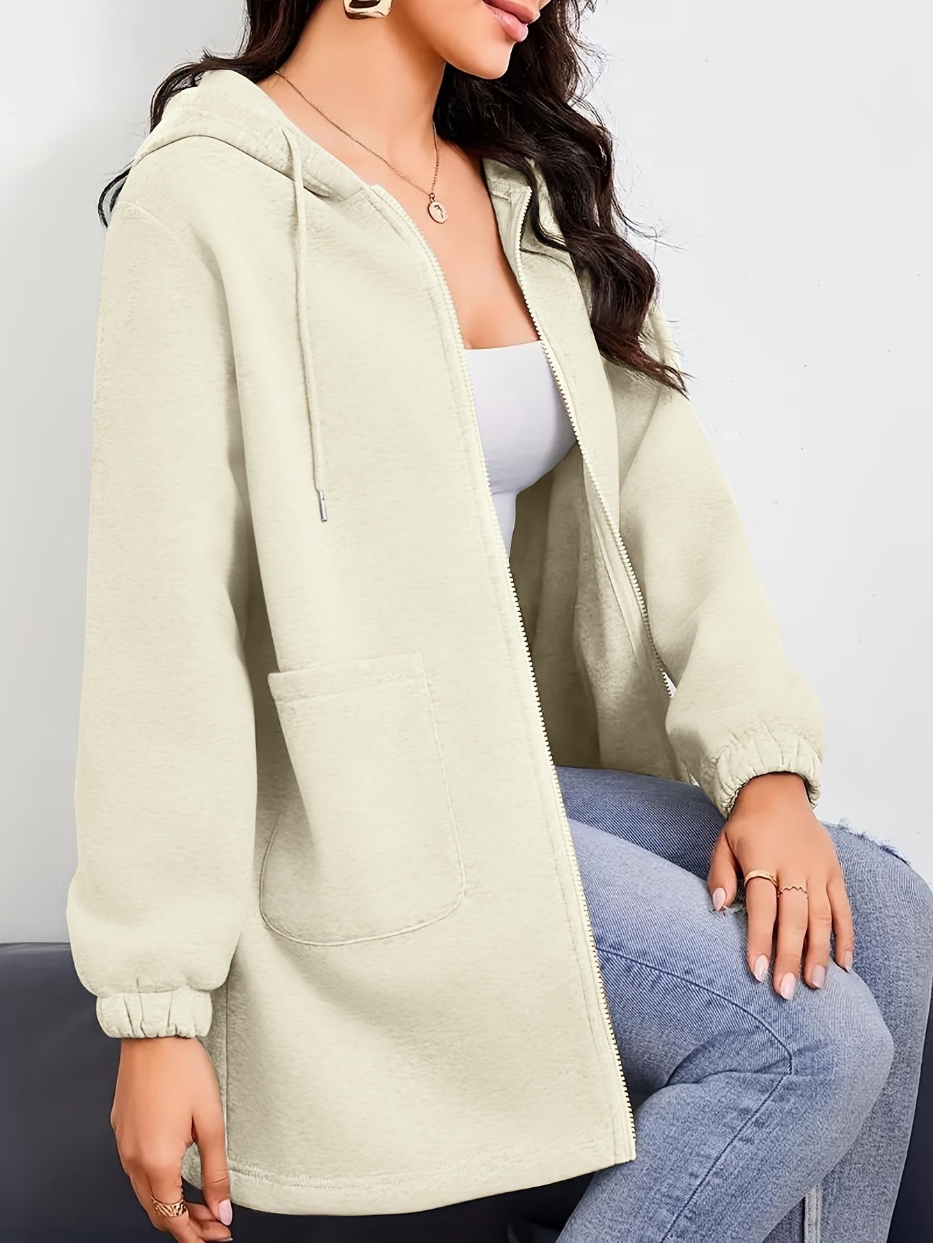 Woman wearing a beige women's oversized zip up hoodies, white crop top, and blue jeans on a plain background