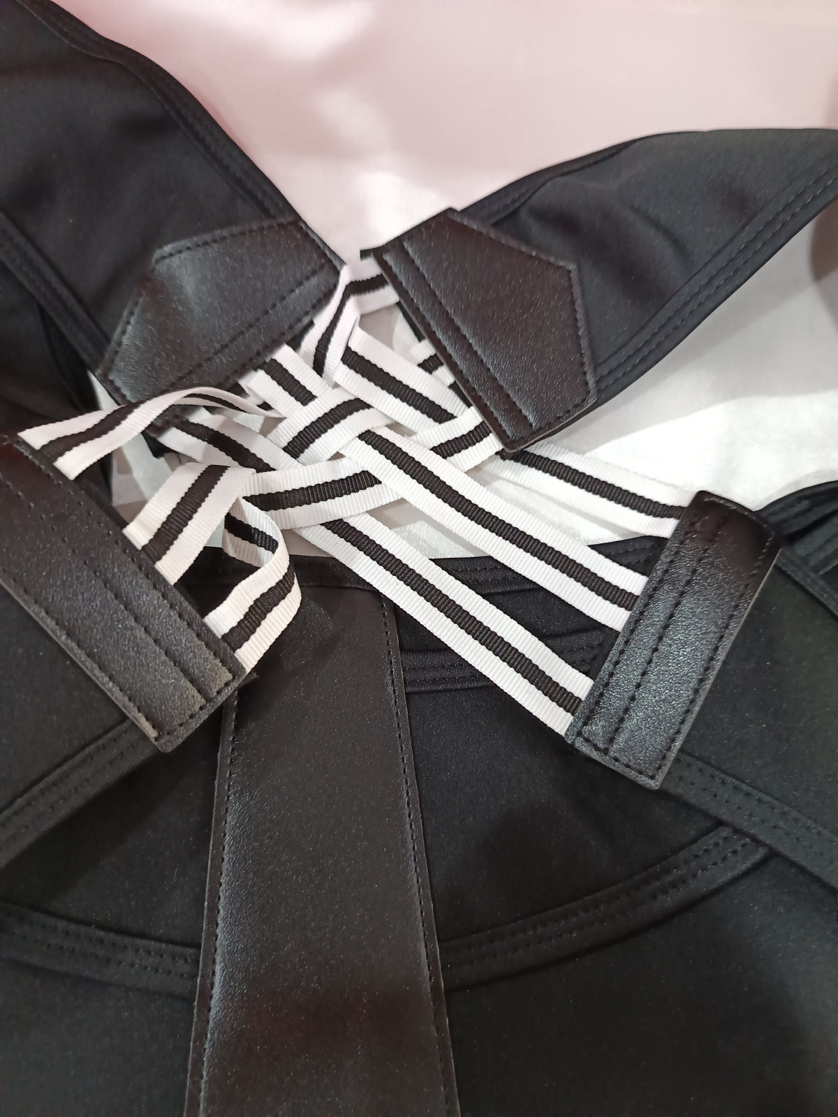 Close-up of a black strappy garment with multiple wide black-and-white striped elastic bands crossing at the back, photographed against a pale pink background.