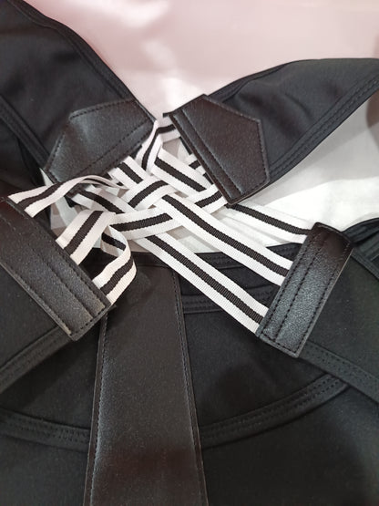 Close-up of a black strappy garment with multiple wide black-and-white striped elastic bands crossing at the back, photographed against a pale pink background.