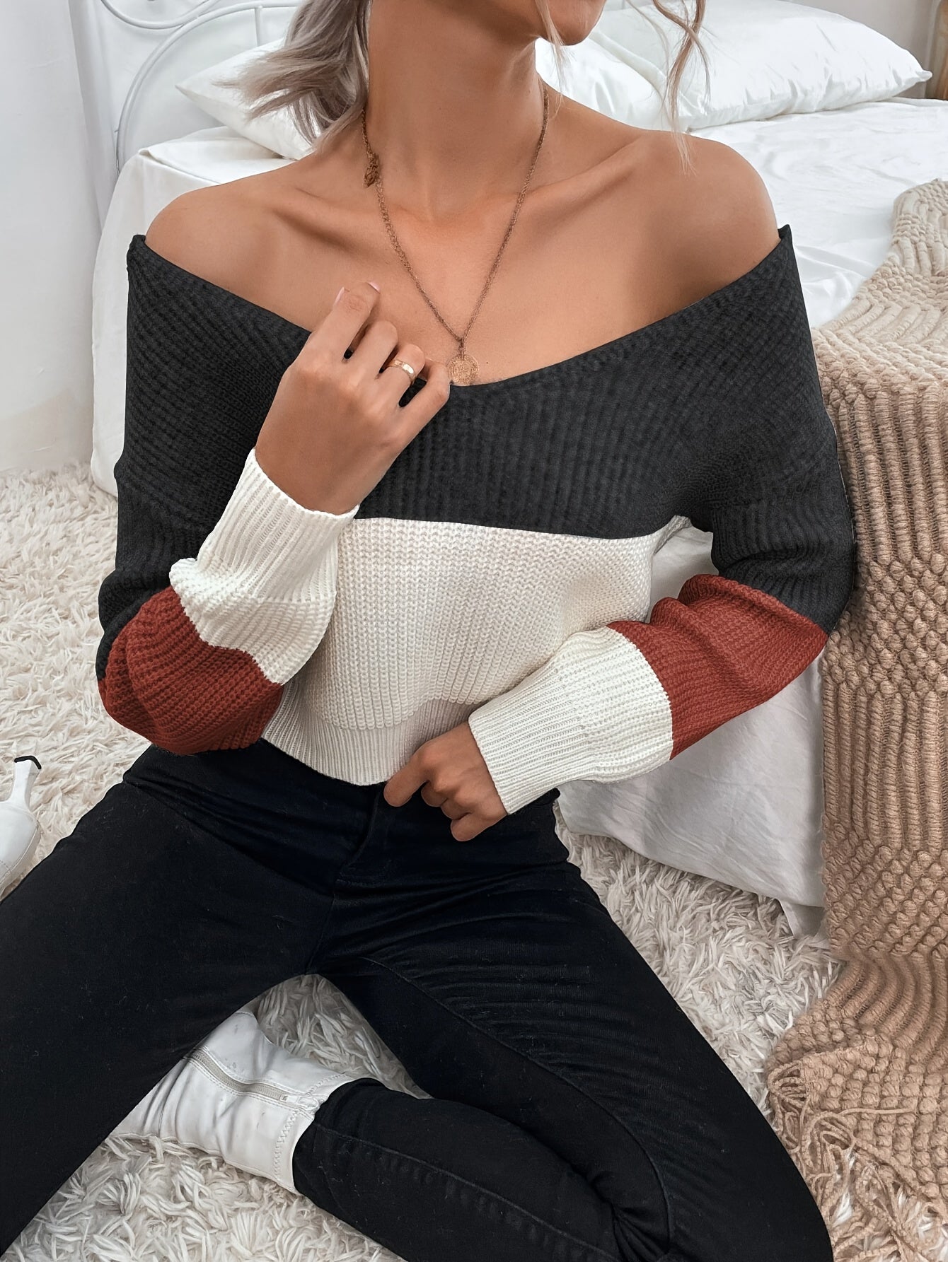 Person wearing a black and white off the shoulder sweater with red sleeves sitting on a bed.