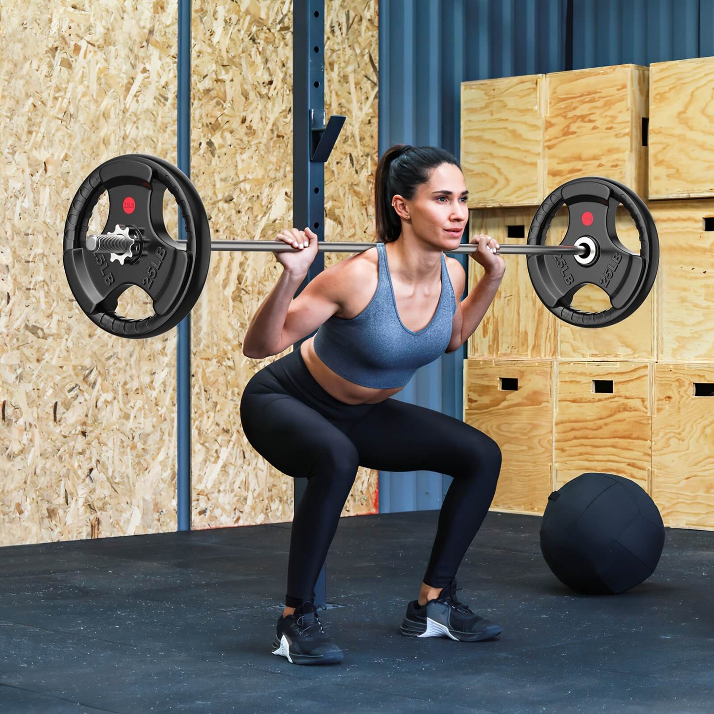Woman lifting a barbell in a gym setting with wooden boxes and a fitness ball in the background.