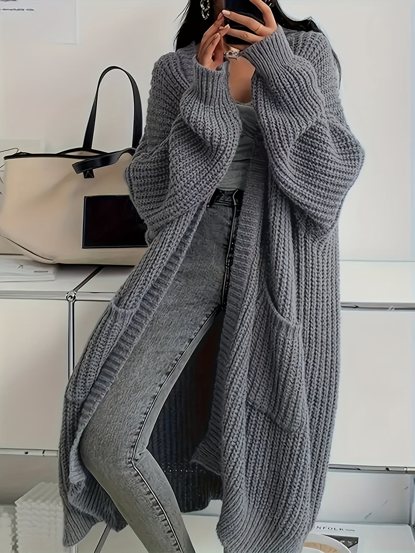 Person wearing a long gray knitted cardigan over jeans, standing against a neutral background.