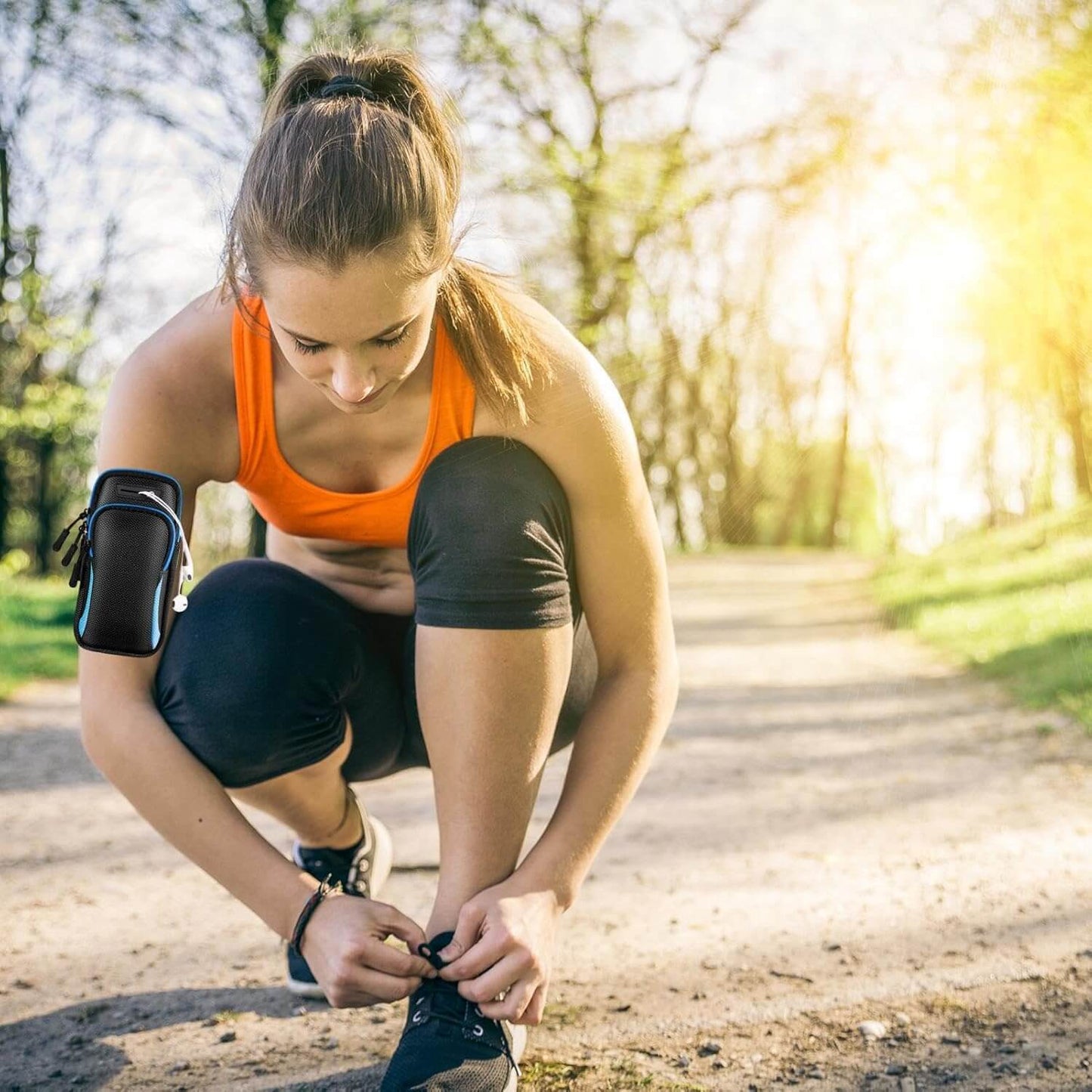 Woman tying her running shoes outdoors on a sunny day while wearing a black arm phone holder for running​ on her fore arm
