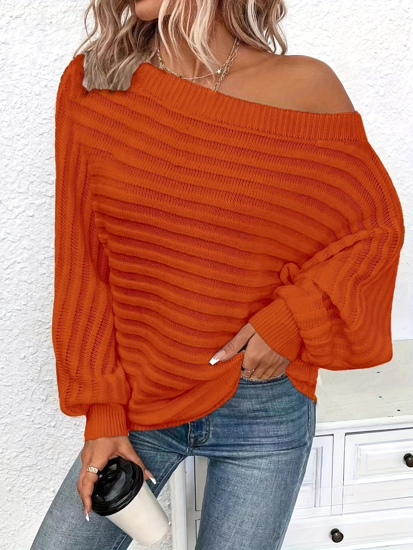 Person wearing an orange off shoulder sweater and blue jeans, holding a coffee cup.