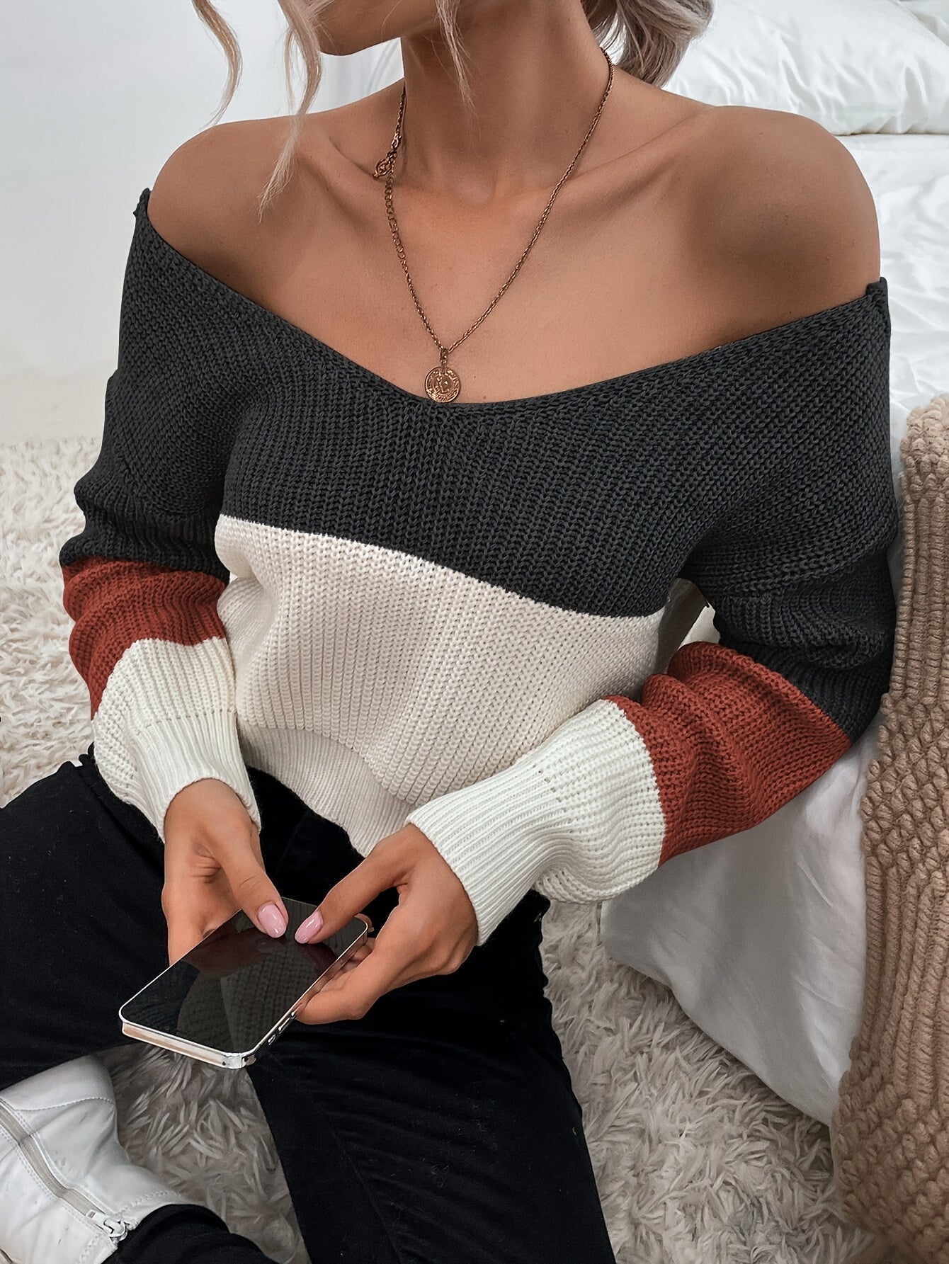 Person wearing a black, white, and brown color-blocked off the shoulder sweater holding a phone.