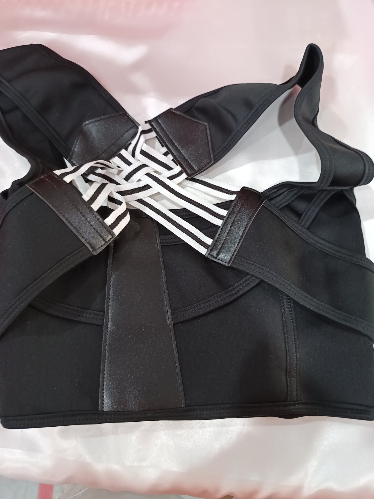 Black strappy support bra with faux-leather straps and a white-and-black striped center band, laid flat on a light pink satin fabric background.