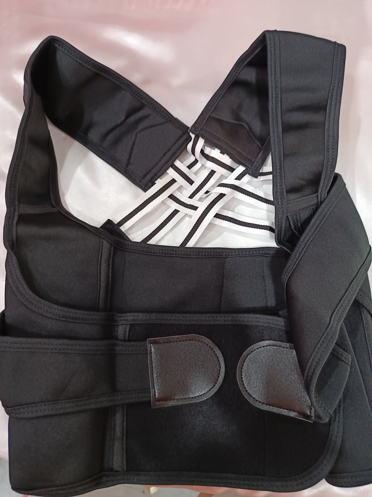 A black posture corrector brace with adjustable straps and hook-and-loop closures, laid flat on a light pink satin fabric background.