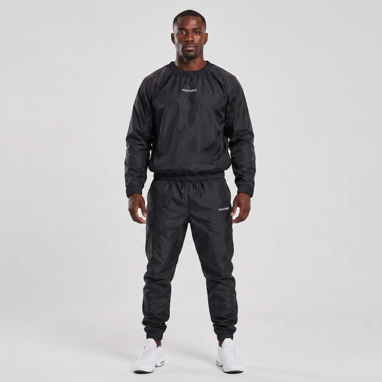 A man modeling a black sauna suit, featuring a long-sleeve windbreaker top and matching jogger pants, paired with white sneakers, against a light gray studio background.