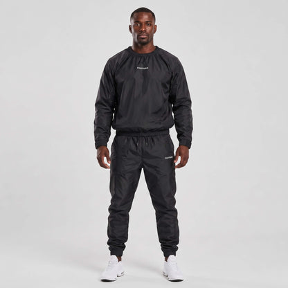 A man modeling a black sauna suit, featuring a long-sleeve windbreaker top and matching jogger pants, paired with white sneakers, against a light gray studio background.