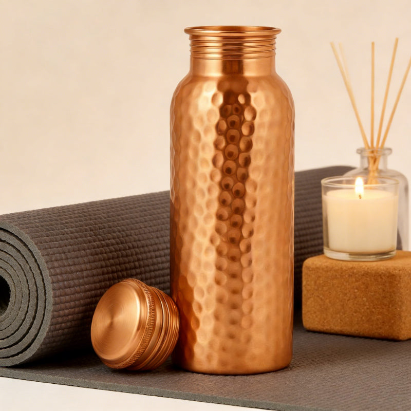Copper water bottle with textured design on a yoga mat with candle and reed diffuser in the background