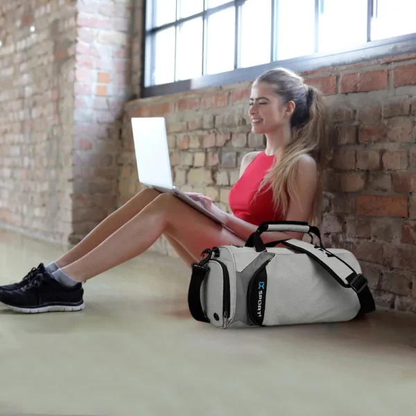 A lady wearing sporty clothes, sitting on the floor while working with her laptop with a workout bag sold by hundred spartans right next to her