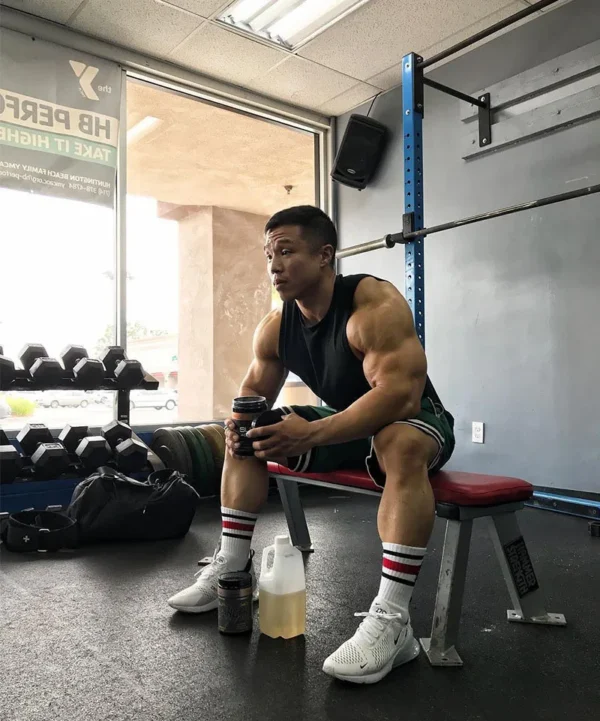 A body builder sitting down and drinking something from a cup while wearing a mens tank top and gym shorts sold by 100 Spartans